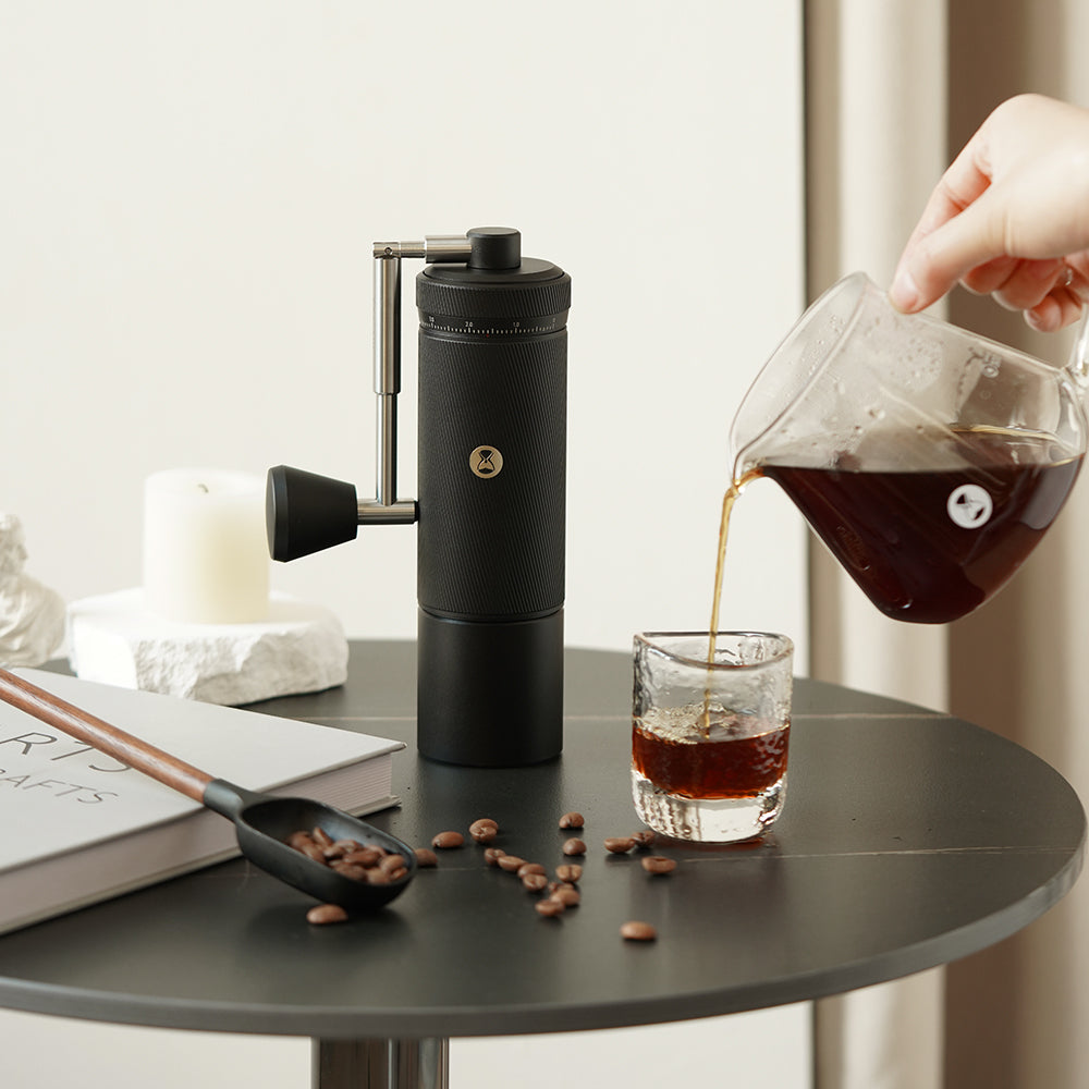 Hand pouring coffee from a glass carafe into a glass on a table with a coffee grinder and coffee beans.