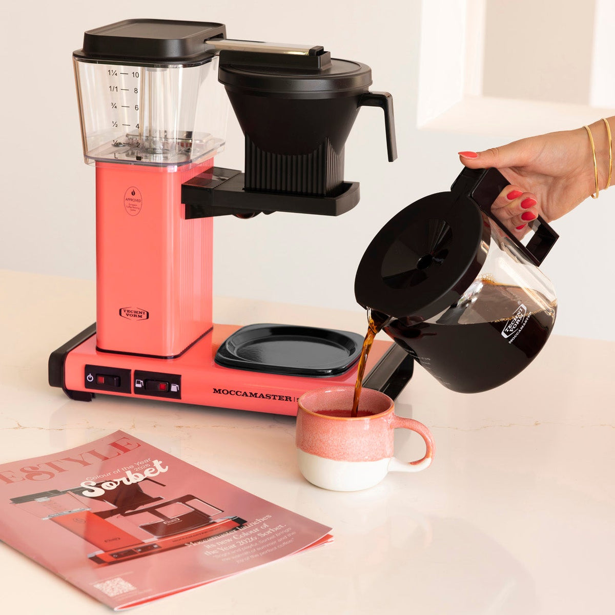 A pink and black coffee maker sits on a white countertop with a person pouring brewed coffee from a glass carafe into a pink and white mug. A coffee magazine lies on the counter nearby.